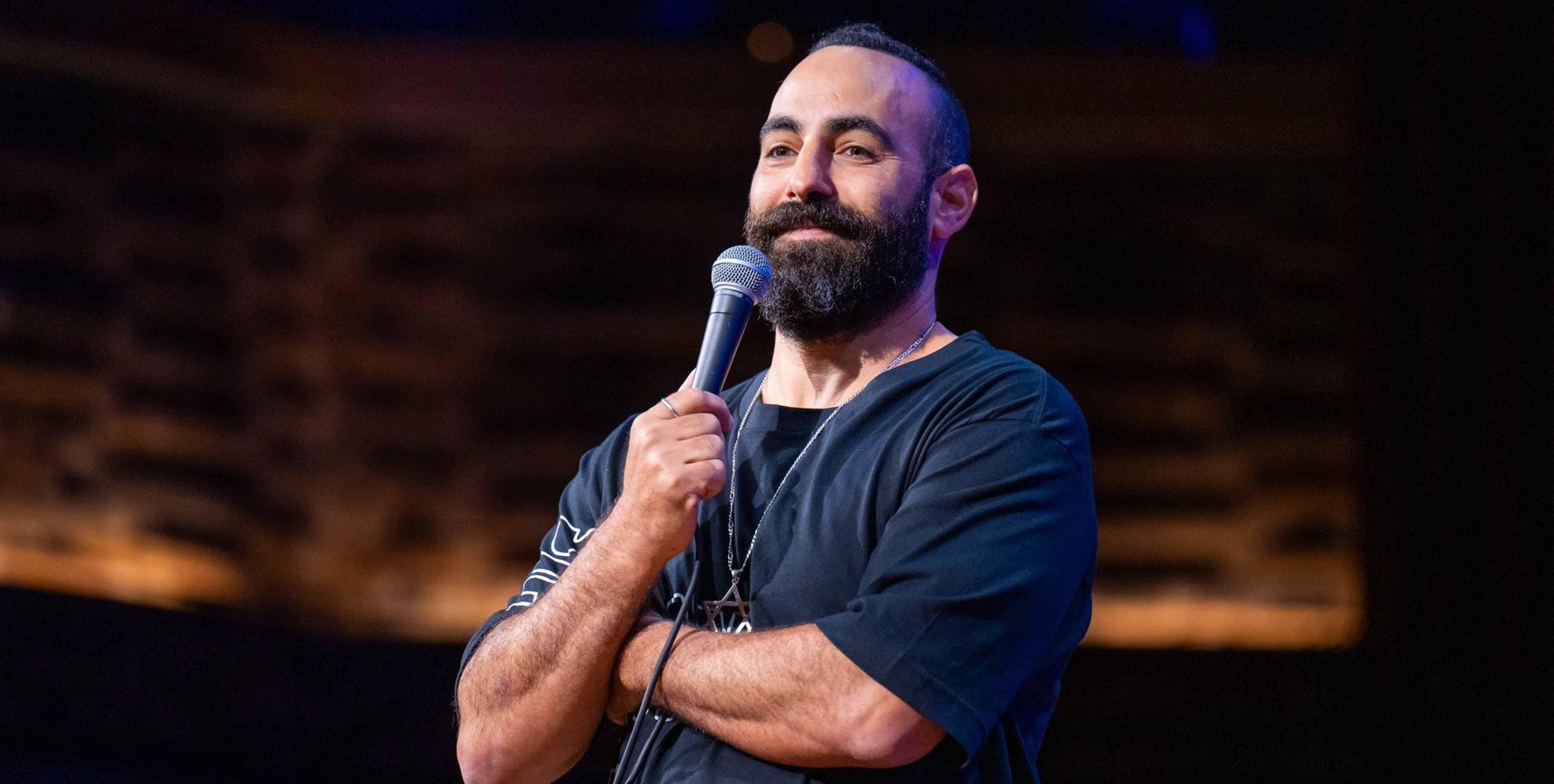 Yohay Sponder on a darkened stage, arms folded, waiting out a laugh